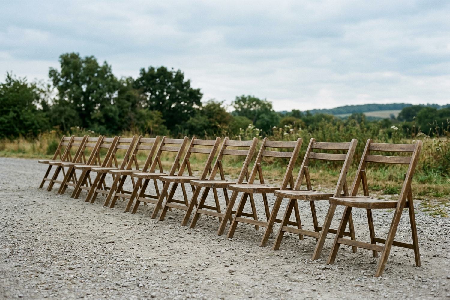 A single row of about twelve empty wooden chairs on gravel under open sky