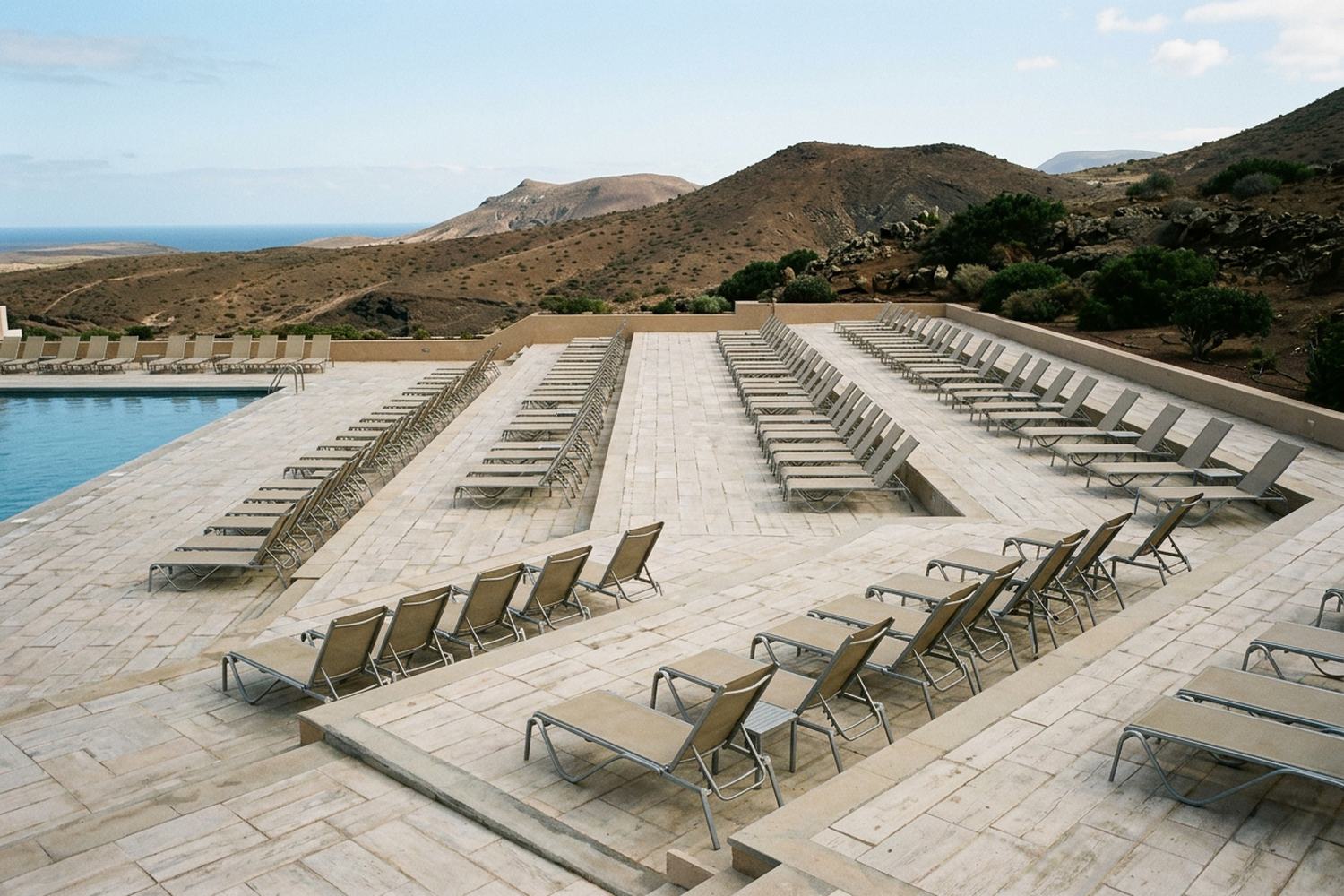 A very large empty terraced pool deck of pale tiles with rows of empty loungers