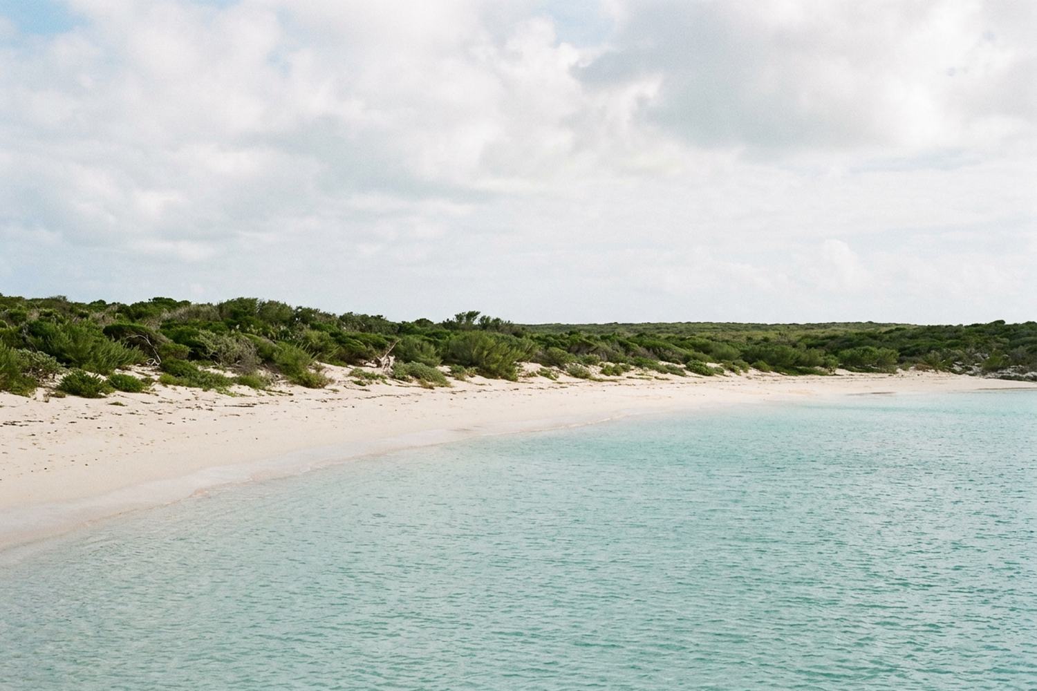 A long shallow turquoise bay with a wide pale sand beach and low green scrub