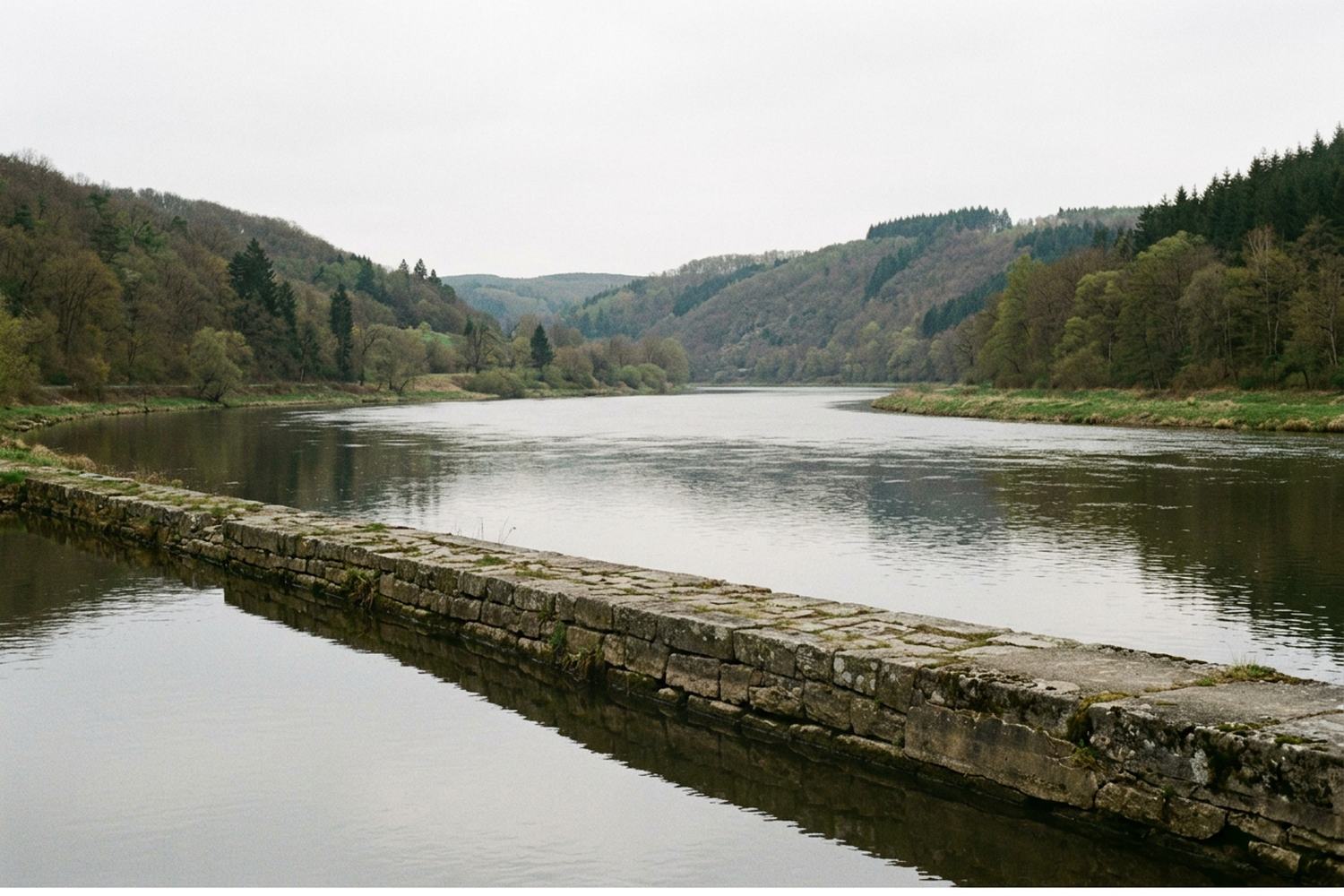 A broad calm river between wooded hills with a low stone embankment
